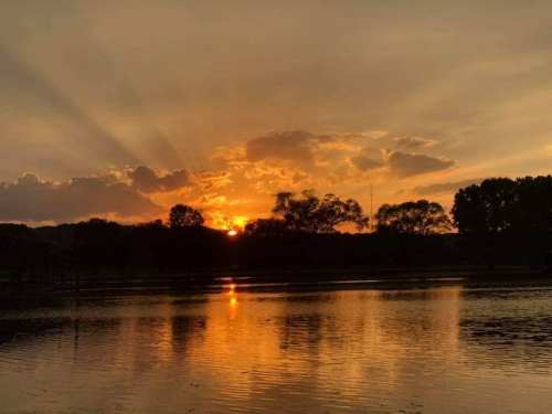 A serene sunset over a lake, with vibrant orange and yellow hues reflecting on the water and silhouetted trees.