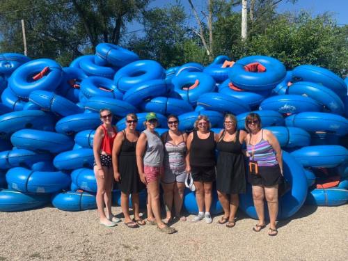 A group of seven women stands in front of a large stack of blue inflatable tubes on a sunny day.