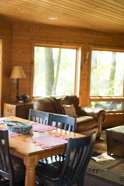 Cozy wooden cabin interior with a dining table, chairs, and a leather sofa by large windows overlooking trees.