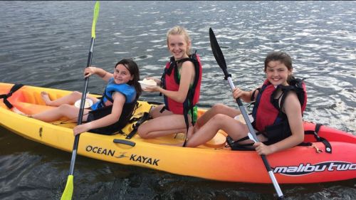 Three girls in a kayak on water, smiling and enjoying snacks, wearing life jackets.