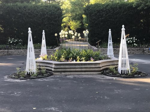 A landscaped garden with white trellises, surrounded by flowers and greenery, leading to a central circular arrangement.