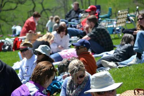 A group of people enjoying a sunny day outdoors, sitting on blankets in a grassy area, some wearing hats and sunglasses.