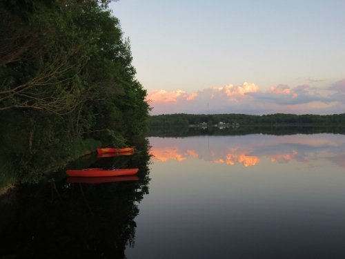Serene lake at sunset with orange kayaks and colorful clouds reflected in the calm water. Lush greenery along the shore.