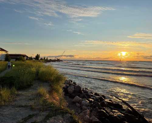 A serene beach scene at sunset, with gentle waves, grassy shoreline, and a distant silhouette of a building.