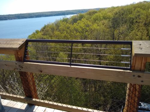 A wooden railing with metal bars overlooks a lush green landscape and a calm lake in the distance.