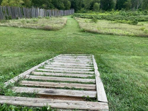 A wooden ramp leads down to a grassy path, surrounded by fields and trees in a serene outdoor setting.