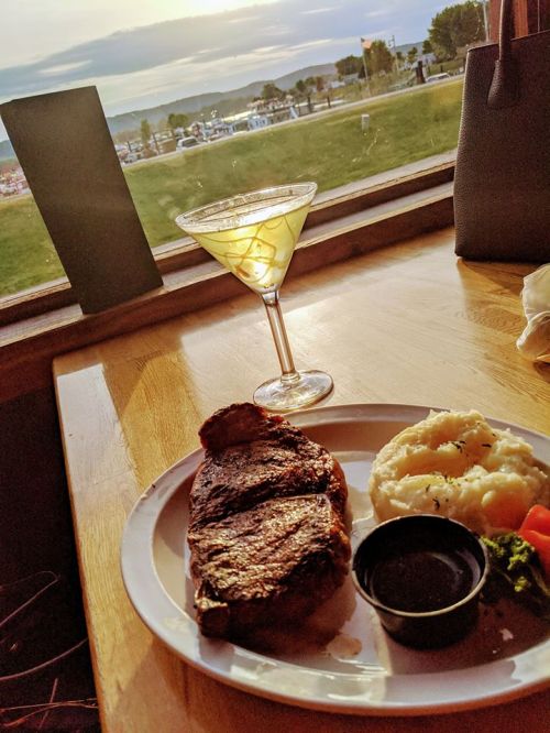 A plate with a grilled steak, mashed potatoes, and a cocktail on a wooden table, with a scenic view outside.