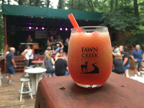 A cocktail in a glass with "Fawn Creek" logo, set against a lively outdoor music venue with people enjoying the performance.