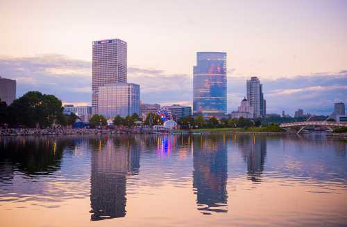 City skyline at dusk, reflecting in calm water, with colorful lights and buildings against a cloudy sky.