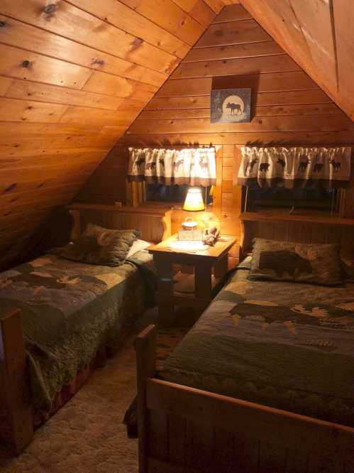 Cozy attic bedroom with two wooden beds, warm lighting, and rustic decor featuring wooden walls and patterned quilts.