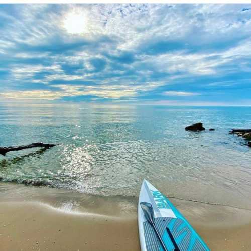 A calm beach scene with a paddleboard on the sand, gentle waves, and a cloudy sky reflecting on the water.