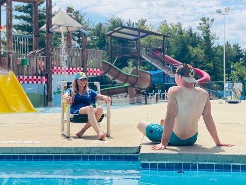 A woman in a blue shirt sits in a chair by the pool, chatting with a shirtless man sitting on the edge. Water slides in background.