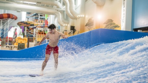 A young man in red shorts surfing on a wave at an indoor water park, splashing water around him.