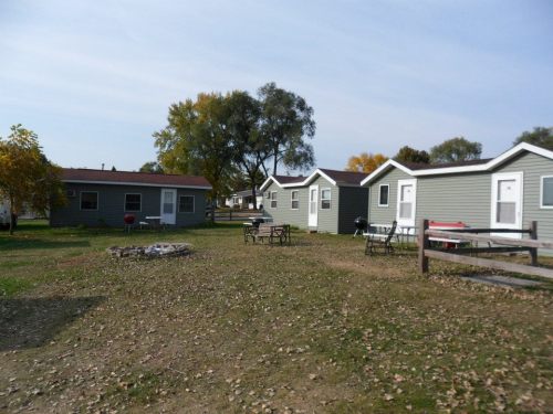 A grassy area with several small cabins, picnic tables, and a fire pit surrounded by trees under a clear sky.