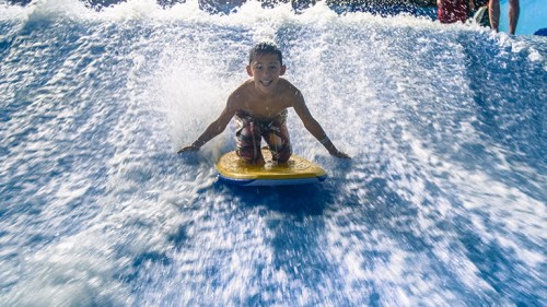 A young boy rides a boogie board on a wave, splashing water around him, with a joyful expression on his face.