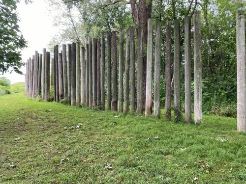 A row of tall wooden posts stands along a grassy area, surrounded by trees and greenery.