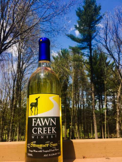 A bottle of Fawn Creek Winery's "Summer Sun" wine against a backdrop of trees and a blue sky.