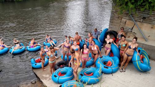 A group of people in swimsuits gather by a river, surrounded by blue inner tubes, ready for a fun day of tubing.