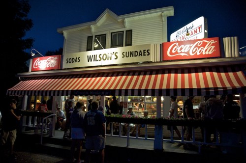 A charming ice cream shop at night, featuring a striped awning and bright Coca-Cola signage, with people outside.
