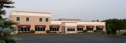 A modern commercial building with large windows and awnings, surrounded by greenery and an empty parking lot.