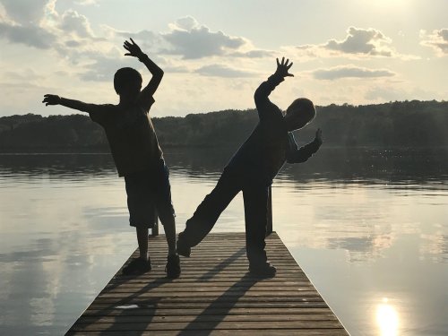 Two children playfully pose on a dock by a calm lake, silhouetted against a sunset sky with clouds.