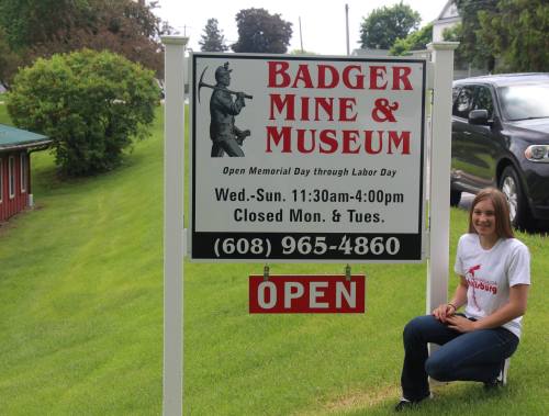 A woman sits beside a sign for Badger Mine & Museum, indicating open hours and contact information.