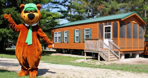 A costumed bear character waves in front of a wooden cabin surrounded by trees.