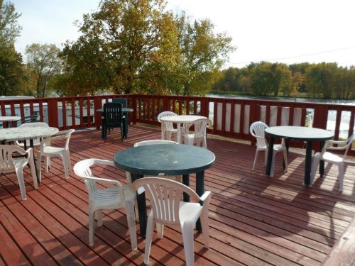 A wooden deck with several tables and chairs, surrounded by trees and a view of water in the background.