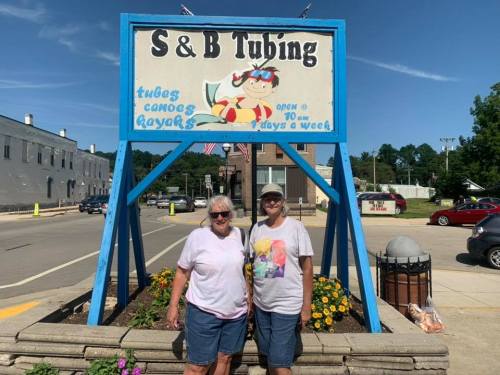 Two women stand in front of a colorful sign for S & B Tubing, featuring tubing and kayaking activities.