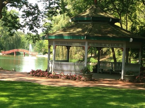 A gazebo by a serene lake, surrounded by lush greenery and colorful flowers, with a red bridge in the background.