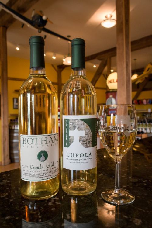 Two bottles of wine labeled "Cupola Gold" and "Cupola," with a wine glass on a dark countertop in a winery setting.