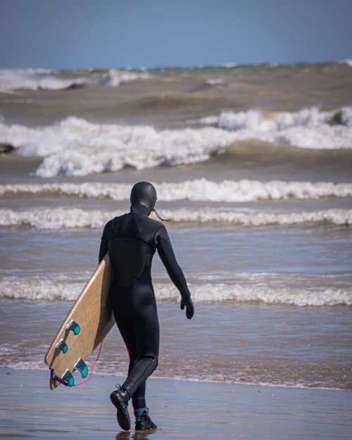 A person in a wetsuit walks along the beach carrying a surfboard, with waves crashing in the background.