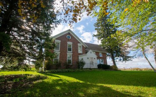 A large brick house surrounded by trees and green grass under a partly cloudy sky.
