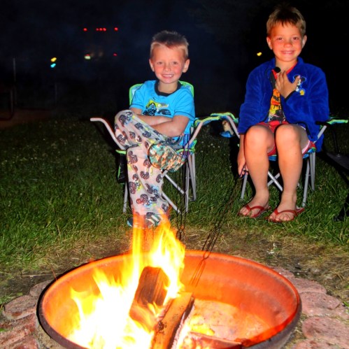 Two young boys sit by a campfire, smiling and enjoying the evening, surrounded by grass and camping chairs.