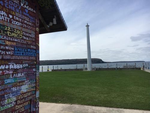 A waterfront view featuring a grassy area, a tall monument, and a colorful wall covered in names and messages.
