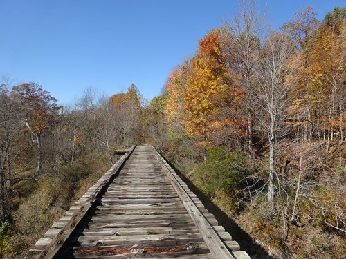 A wooden railway bridge stretches into the distance, surrounded by trees with vibrant autumn foliage under a clear blue sky.