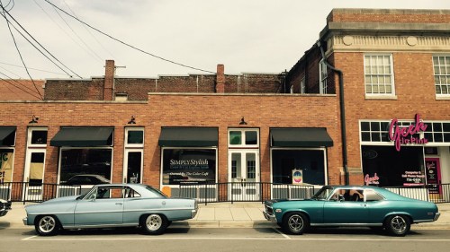 Two vintage cars parked along a street in front of brick buildings with storefronts and awnings.