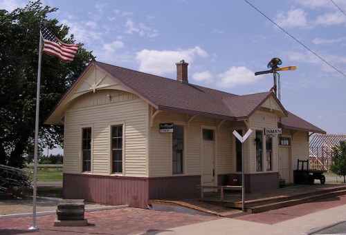 Historic train station with a flag, signal, and platform, set against a blue sky with scattered clouds.