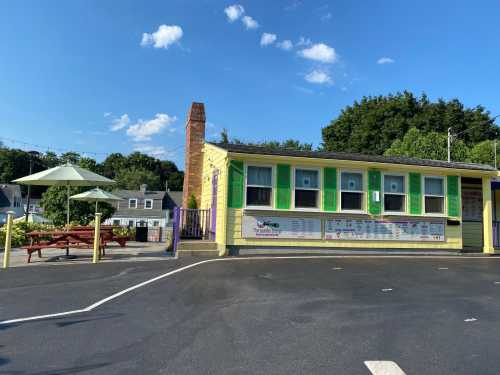 A colorful yellow building with green shutters, picnic tables, and umbrellas under a clear blue sky.