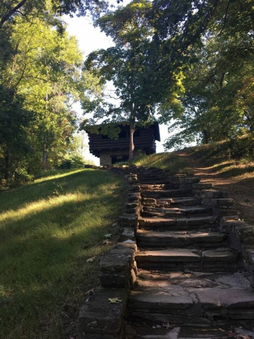 Stone steps lead up a grassy hill to a small building surrounded by trees in a serene outdoor setting.