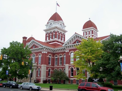 Historic red brick courthouse with a clock tower, surrounded by trees and parked cars under a cloudy sky.