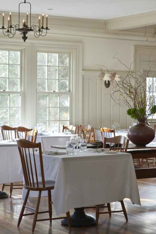 A cozy restaurant interior with wooden chairs, tables covered in white cloths, and large windows letting in natural light.