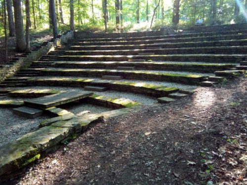 An outdoor amphitheater with stone steps, surrounded by trees and dappled sunlight filtering through the leaves.