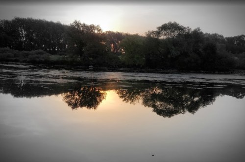 A serene river scene at sunset, with trees reflecting on the calm water's surface.