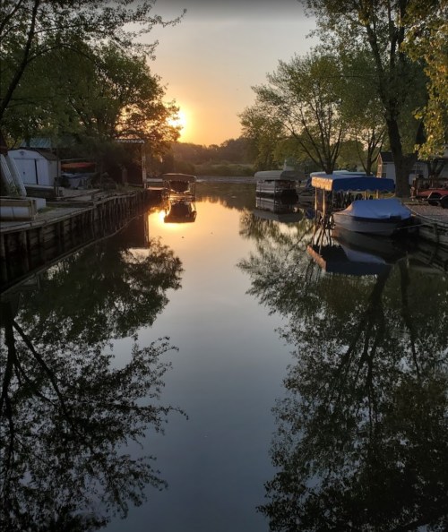 Sunset over a calm canal, with boats and trees reflected in the water, creating a serene atmosphere.