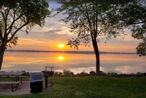 Sunset over a calm lake, with trees framing the scene and a picnic area in the foreground.