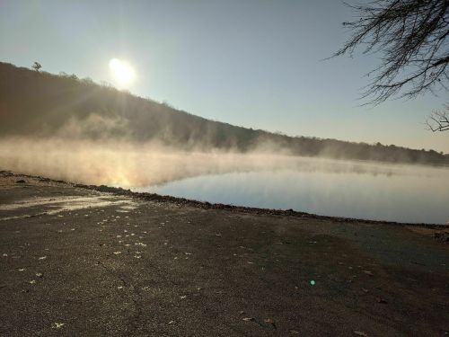 A serene lake at sunrise, with mist rising from the water and a hillside in the background.