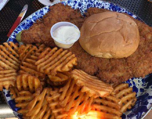 A plate featuring two pieces of fried chicken, a bun, and a side of waffle fries with a small cup of dipping sauce.