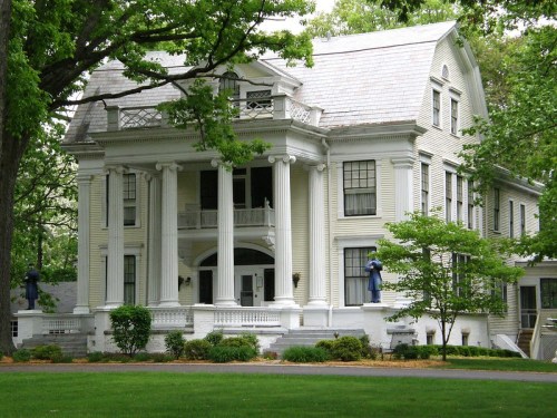 A large, elegant white house with columns, surrounded by trees and greenery, featuring a porch and well-maintained landscaping.