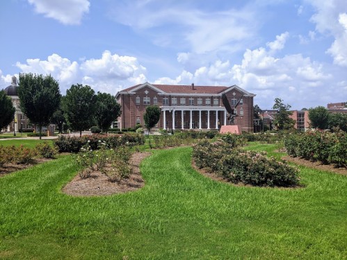 A brick building with columns surrounded by green grass and rose bushes under a blue sky with fluffy clouds.